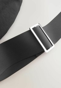 Bolsa de tela negra con una superficie texturizada y un resistente broche rectangular metálico en una correa plana trenzada.