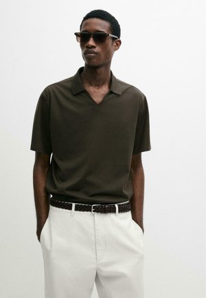 Man draagt een donkergroen polo met korte mouwen, een witte broek, een gevlochten bruine riem en een donkere zonnebril, staat met handen in de zakken tegen een witte achtergrond.