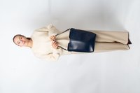 Mujer con un suéter y pantalones beige sosteniendo un gran bolso de cuero azul marino con ambas manos frente a un fondo blanco.