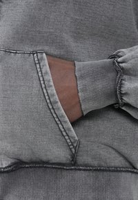 Hanorac gri cu manșete rib și buzunar tip cangur, cu un finisaj texturat. Cusături vizibile de-a lungul marginilor și detaliu de țesătură adunată.