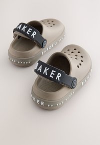 Bež plastične cipele s ventilacijskim otvorima, crni podesivi remen s logotipom "BAKER" i bijelim natpisom duž đona.