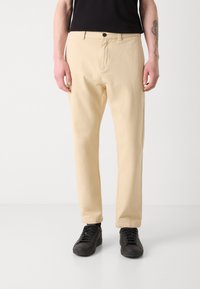 Pantalon en coton beige avec une coupe droite, doté d'une fermeture à bouton à l'avant et de poches latérales, associé à des baskets noires.