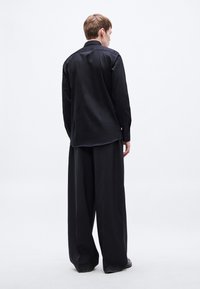 Fekete, hosszú ujjú ing egyenes kivágással, széles fekete nadrággal párosítva. Az anyag simának tűnik, gombos mandzsettával és gallérral.