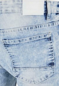 Jeans de mezclilla azul claro con una textura desgastada. Presenta un bolsillo trasero con costuras reforzadas y una etiqueta de tela blanca en la cintura.