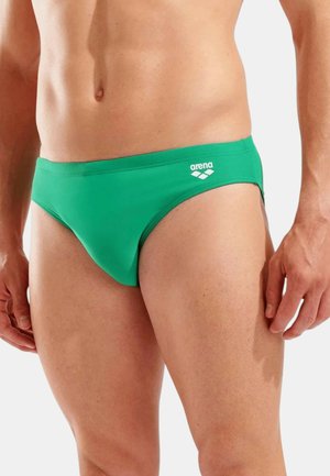 Torace maschile che indossa slip da nuoto verde del marchio Arena, mostrando la zona mediana e la parte superiore delle cosce con le mani rilassate ai lati.