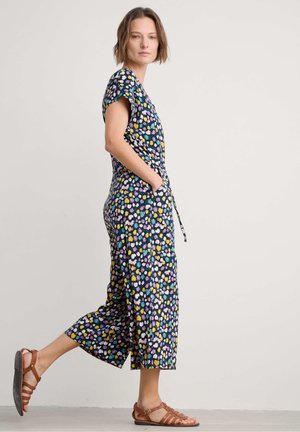 Jumpsuit w kwiatowy wzór w kolorze granatowym, z krótkimi rękawami, wiązanym w pasie i skróconym krojem. Noszony z brązowymi sandałami na paski.