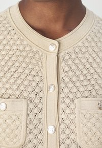 Beige stickad cardigan med ett strukturerat mönster, som har en rund hals, silverknappar och två framfickor.