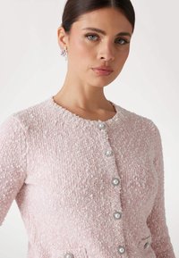 Light pink knitted cardigan with a textured surface, round neckline, and seven pearl-like buttons down the front. No visible pockets.