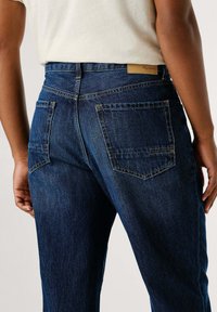 Vaqueros de mezclilla azul oscuro con cintura alta, que presentan dos bolsillos traseros y detalles de costura visibles. La tela tiene una ligera textura.