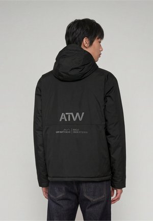 Mann trägt eine schwarze Kapuzenjacke mit reflektierenden "ATW" und "Off Duty Gear" Logos auf dem Rücken, kombiniert mit dunkelblauen Jeans.