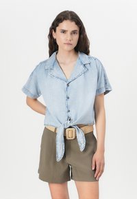 Ανοιχτό μπλε denim πουκάμισο κοντό μανίκι με λεπτομέρεια δέσιμου μπροστά, διαθέτοντας γιακά και κουμπιά, σε συνδυασμό με χακί σορτς και υφαντή ζώνη.