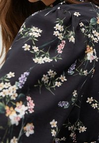 Blouse en tissu noir avec des manches bouffantes, ornée d'un motif floral en blanc, rose et lavande. Texture douce avec des fronces le long de l'encolure.