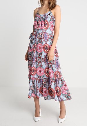 Kobieta ubrana w sukienkę bez rękawów sięgającą kostek, z czerwono-niebiesko-białym geometrycznym wzorem kwiatowym, oraz białe buty na wysokim obcasie.