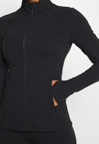 Černá sportovní bunda s vysokým límcem, zapínáním na zip, přiléhavým střihem a bočními kapsami. Hladká, pružná látka s plochými švy.