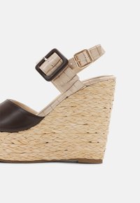 Sandale compensée en beige tissé avec bride en cuir marron foncé sur les orteils et bride beige à la cheville avec boucles dorées et noires.