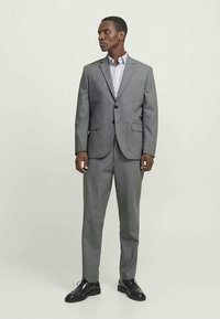 Homem vestindo um fato cinza estampado com sapatos pretos e uma camisa de botão listrada, de pé contra um fundo claro, olhando para a esquerda.