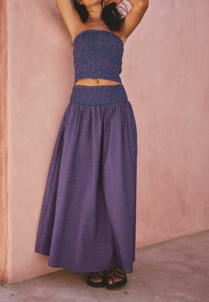 Frau, die ein trägerloses, strukturiertes blaues Crop-Top und einen passenden langen Rock trägt, barfuß vor einer hellrosa Wand stehend.