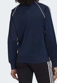 Osoba v tmavě modré bundě s dlouhým rukávem a bílým lemem na ramenou, a černých kalhotách se třemi bílými pruhy po stranách.