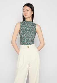 Groene bloemenmouwloze top met een hoge hals, gekenmerkt door een patronenontwerp en een gladde textuur, gecombineerd met hoog opgesneden crèmekleurige broeken.