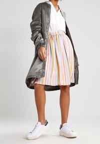 Femme portant un manteau gris brillant sur un haut blanc, une jupe rayée pastel et des baskets blanches, se tenant devant un fond uni.