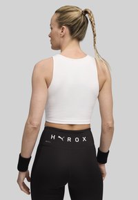 Witte geribbelde cropped top en zwarte high-waisted leggings met "HYROX" opdruk. Zwarte polsbanden. Glad textuur, aansluitend design.