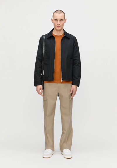 Sort letvægtsjakke med lynlås foran, orange strikket skjorte, beige cargo-bukser og hvide sneakers. Sort skuldertaske med rem.