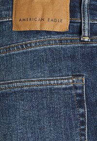 Sininen farkkulegginsit, joissa on teksturoitu pinta, joka on varustettu erottuvalla keltaisella ompeleella ja ruskealla nahkalapulla, jossa on kohokuvioitu "AMERICAN EAGLE".