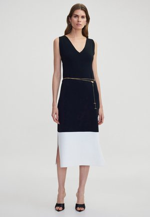 Abito senza maniche in bianco e nero con scollo a V, caratterizzato da una cintura in catena dorata, uno spacco laterale e una superficie liscia. Il design è contemporaneo e allungato.
