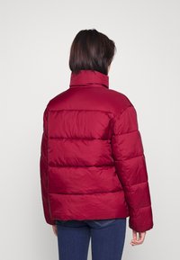 Rød puffjakke med høy krage, med en quiltet design og en glatt tekstur. Baksiden viser en boksformet silhuett og lange ermer.