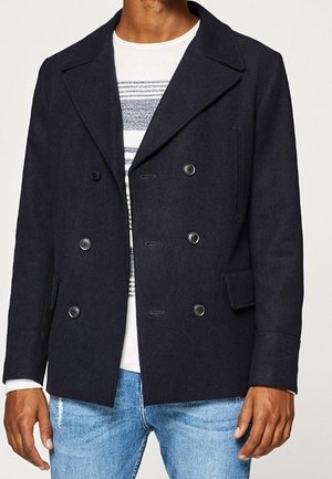 Mężczyzna w ciemnogranatowym dwurzędowym płaszczu typu pea coat, białej koszuli w paski i jasnoniebieskich dżinsach, stojący na jednolitym tle.