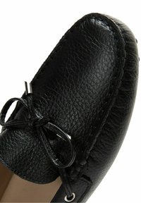 Mocasín de cuero negro con textura granulada, con un lazo en la parte frontal, detalles plateados y costuras de contraste a lo largo de los bordes.