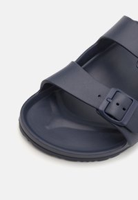 Chausson en toile bleu marine avec deux larges sangles à surface texturée et une boucle réglable sur le côté. Matériau durable et flexible.