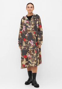 Kurtka przeciwdeszczowa w kamuflażowy wzór z kwiatowymi akcentami w ziemistych odcieniach; posiada kaptur, długie rękawy i luźny krój. Noszona z czarnymi butami.