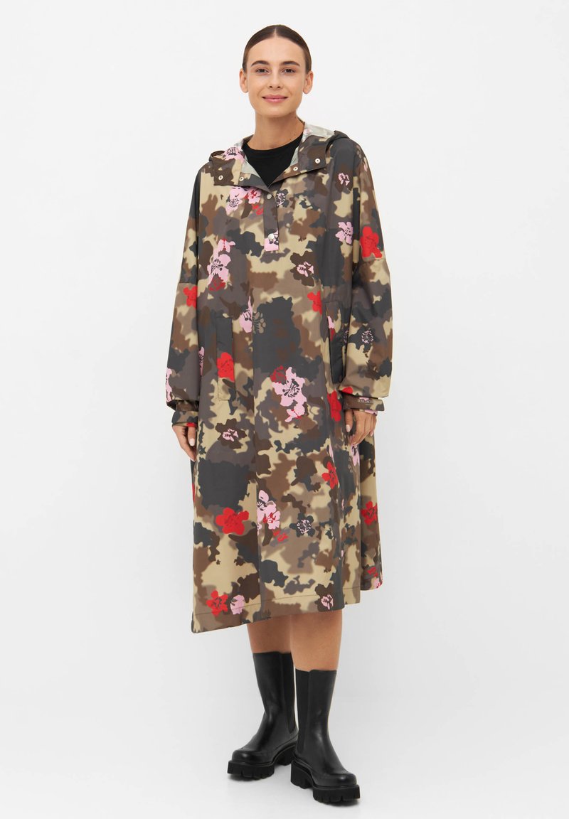 Kurtka przeciwdeszczowa w kamuflażowy wzór z kwiatowymi akcentami w ziemistych odcieniach; posiada kaptur, długie rękawy i luźny krój. Noszona z czarnymi butami.