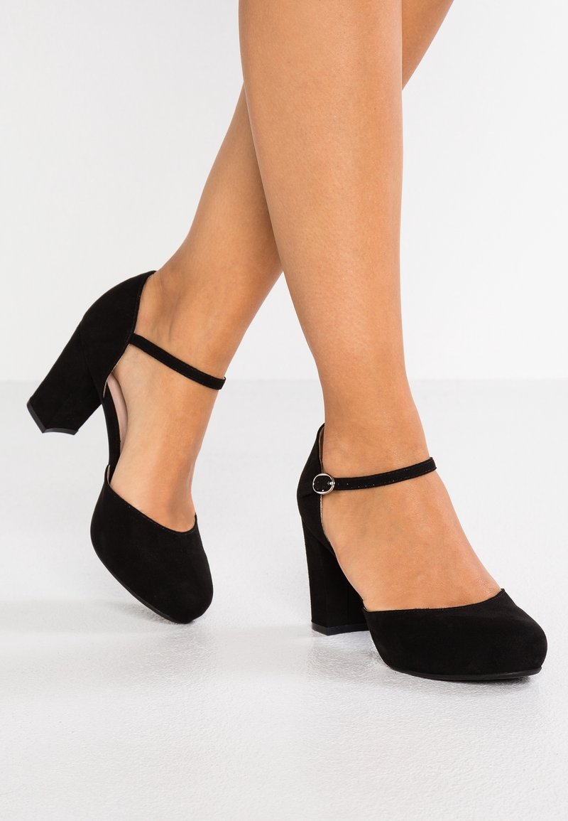 Jambes portant des escarpins Mary Jane noirs à bouts fermés avec des brides autour de la cheville et des talons blocs arrondis, sur un sol blanc.