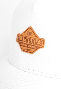 Hvidt kasket med en brun læderlap, der viser "BLACKSKIES PREMIUM SUPPLIES MMIX." Har tekstureret stof og syningsdetaljer.