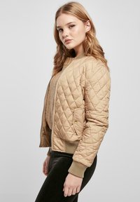 Jeune femme aux cheveux blonds portant une veste bomber beige matelassée sur un pull en tricot, pantalon noir, debout de profil sur fond gris.