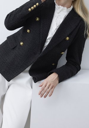 Frau trägt einen strukturierten schwarzen zweireihigen Blazer mit goldenen Knöpfen und eine weiße Bluse mit Rüschen, lehnt sich auf eine weiße Fläche, weiße Hose sichtbar.