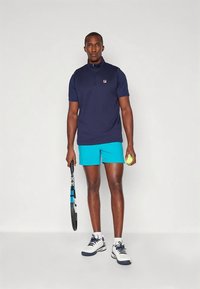 Polo shirt blu navy con colletto con zip e logo del marchio; pantaloni corti blu brillante; tiene una racchetta da tennis e una pallina; indossa scarpe da ginnastica bianche e blu navy.