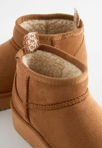 Brune ankelstøvler med en suede yderside, blød falsk pelsforing og en vævet detalje bagpå. Har en flad, tekstureret sål.