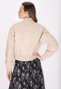 Donna con lunghi capelli castani che indossa una giacca beige con maniche e orlo a costine, abbinata a una gonna nera con un motivo floreale bianco.