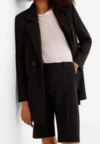 Blazer noir à rayures avec un col peg, porté sur un t-shirt blanc, associé à un short à rayures assorti. Texture de tissu lisse.