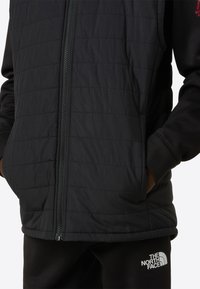 Čierna prešívaná vesta s predným zipsom, zdobená jemnými švami. Kombinovaná s čiernym svetríkom a teplákmi s logom.