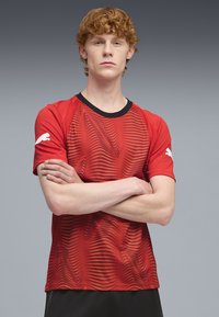 Camiseta deportiva roja con patrones de ondas, textura acanalada y detalles en el cuello de color negro. Logo blanco en la manga izquierda. Diseño de manga corta.