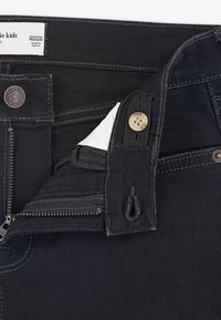 Pantalones de mezclilla negra con cremallera en la bragueta, cierre de botón y pasadores para cinturón. La tela tiene una textura suave con costuras y etiquetas en contraste.