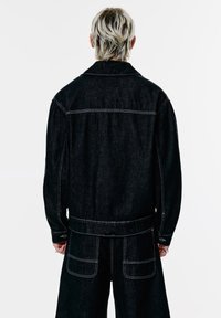 Person med kort, lagdelt blond hår iført en sort denimjakke og matchende brede bukser, set fra ryggen.