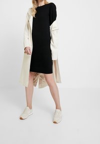 Robe noire en maille avec un col rond, associée à un long manteau beige clair. Des baskets blanches avec des semelles texturées complètent la tenue.