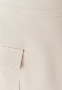 Beige stof met een gladde textuur, voorzien van een discreet pat pocket en subtiele stikdetails. Minimalistisch ontwerp en een zachte uitstraling.