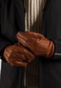 Bruine leren handschoenen met gestikte details, een aangesloten ontwerp en een gladde textuur. Draagt door een persoon met een gebreide trui en een donker jack.