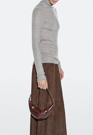 Frau in grauem Rollkragenpullover und braunem Rock hält eine kleine, glänzende dunkelbraune Ledertasche am Henkel.